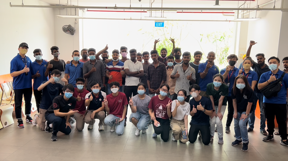 A large group of people wearing face masks pose together indoors, many giving thumbs up and peace signs.